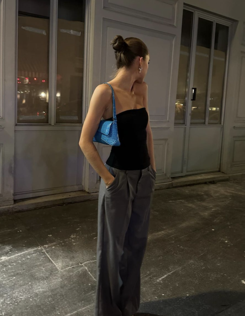 Woman in an elegant evening outfit with a black strapless top, grey trousers, pointed silver heels, and a blue shoulder bag, standing on a city sidewalk.